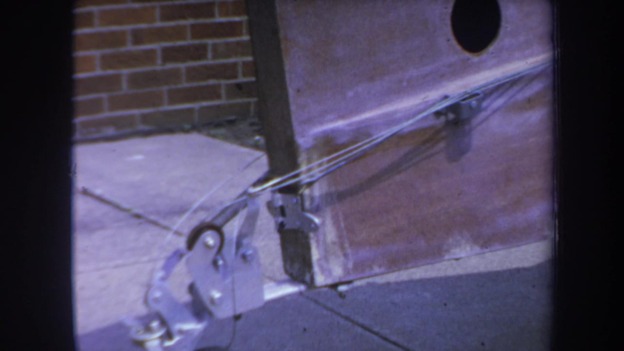 1969 Yucaipa-area 8mm frame showing a close mechanical trolley detail with taut wires, metal brackets, and a small wheel near concrete.
