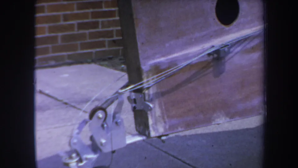 1969 Yucaipa-area 8mm frame showing a close mechanical trolley detail with taut wires, metal brackets, and a small wheel near concrete.