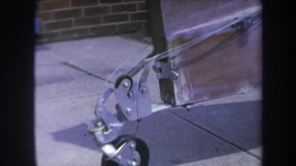1969 8mm frame showing the lower trolley wheel and riveted metal armature against sidewalk, brick, and a reddish panel.
