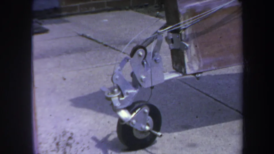 Close final frame of the 1969 Yucaipa trolley detail, showing the wheel, bracket, taut wires, sidewalk, and adjacent metal panel.
