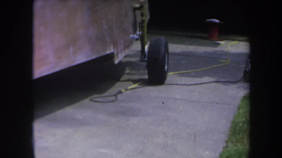 1969 Yucaipa frame showing the homemade trailer from the side, with a wheel, yellow cord, concrete driveway, and grass edge.