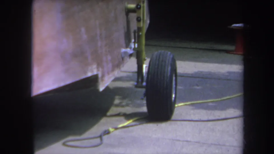 Close 1969 8mm view of the trailer tire and yellow-painted hardware, with a driveway cord and dark garage area behind it.
