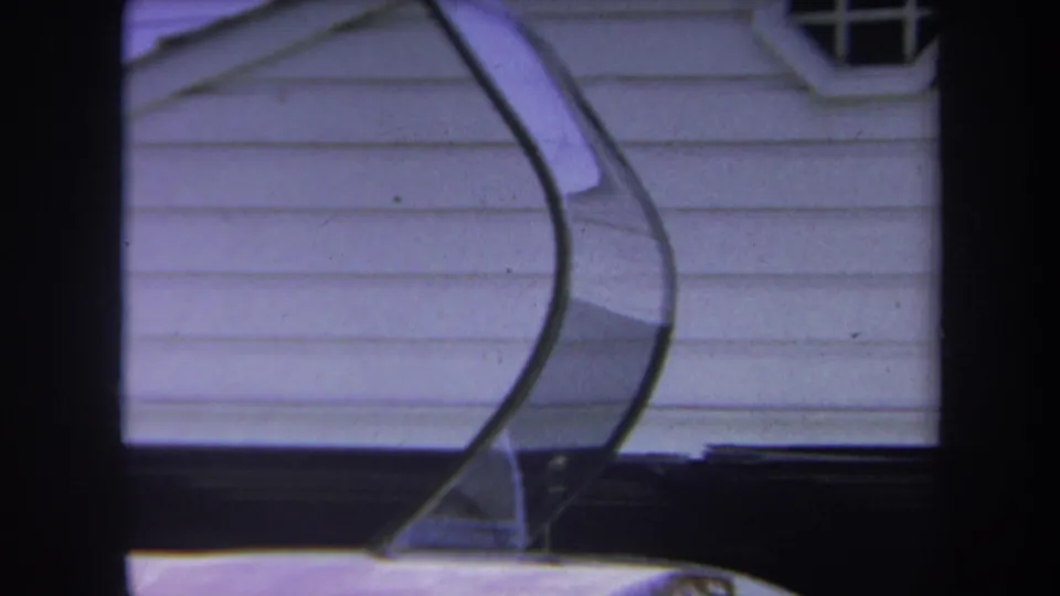 1969 home-movie frame with the helicopter's curved canopy and windshield frame beside a white house wall.