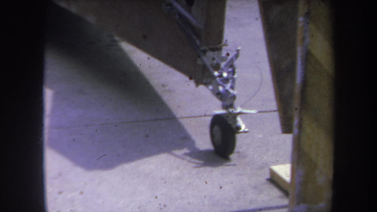 1969 Yucaipa-area 8mm frame showing part of a gray aircraft component with a small wheel on concrete under a shelter.