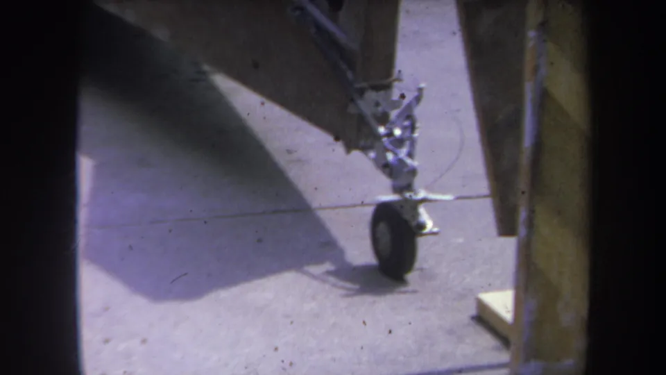 1969 Yucaipa-area 8mm frame showing part of a gray aircraft component with a small wheel on concrete under a shelter.
