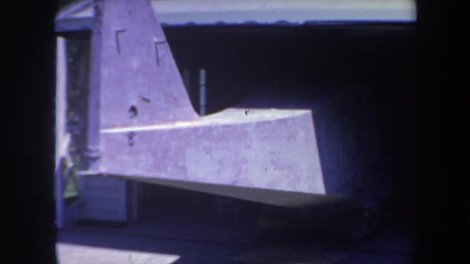 1969 Yucaipa-area home-movie frame showing the side of a gray aircraft part in shade under a simple shelter.