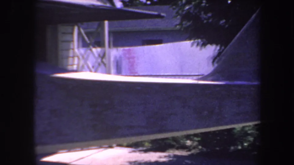 Close 8mm view of the aircraft tail area near a house, with a ladder, shaded trees, and bright sun patches in the yard.