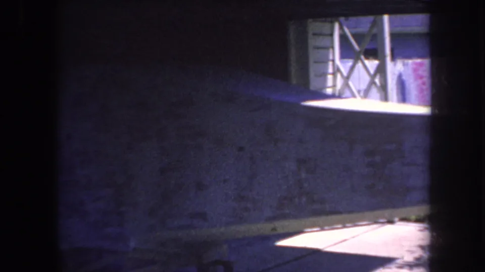 1969 Yucaipa-area home-movie frame with a dark foreground, textured wall, wood trim, cross-bracing, and sunlit floor shape.