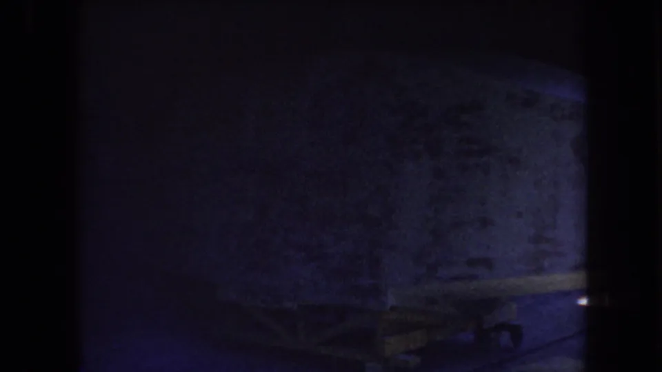 Very dark 1969 Yucaipa-area 8mm frame with a textured wall, faint lower structural shapes, and strong purple-blue exposure shift.