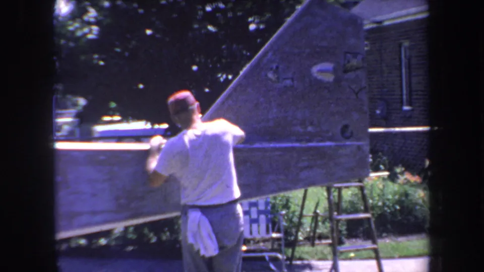 The man continues working on the aircraft tail section outdoors, with ladder, plants, and building exterior visible in 1969 California footage.
