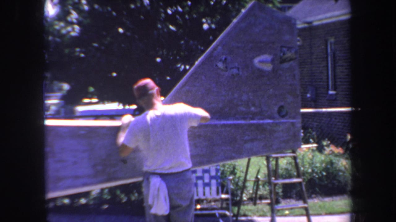 1969 Yucaipa 8mm frame of a man seen from behind sanding a large wooden hull-shaped structure near a house and ladder.