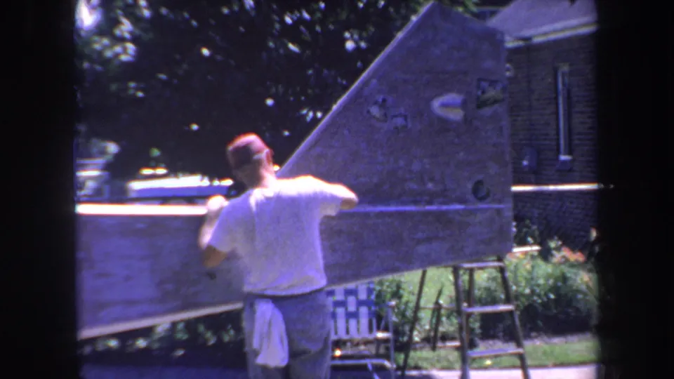 1969 Yucaipa 8mm frame of a man seen from behind sanding a large wooden hull-shaped structure near a house and ladder.