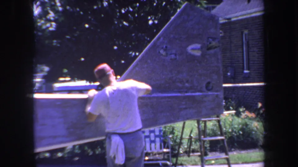 The man works along the side of the wooden hull in the backyard, with a ladder, brick wall, greenery, and strong sunlight visible.