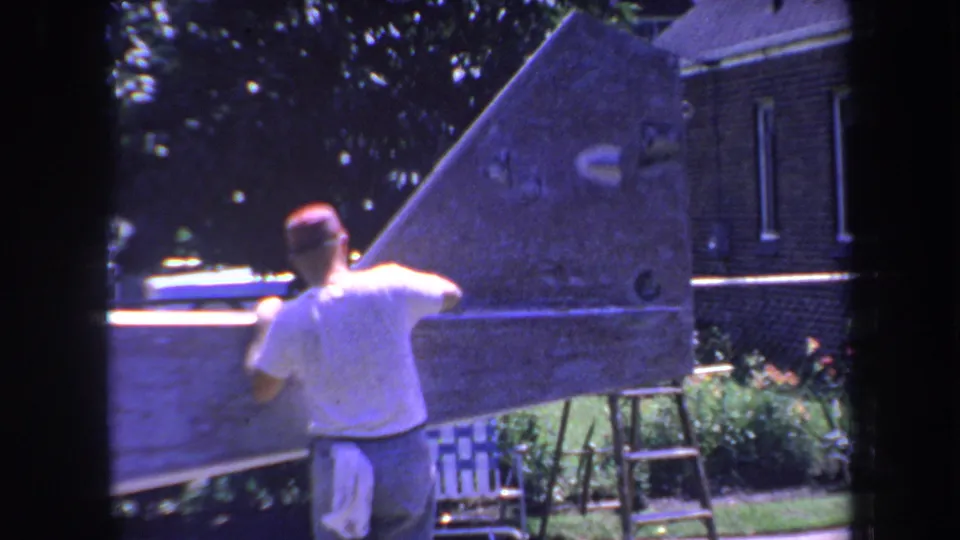 A handheld 8mm view shows the man from behind as he sands the broad wooden surface in a California backyard setting.