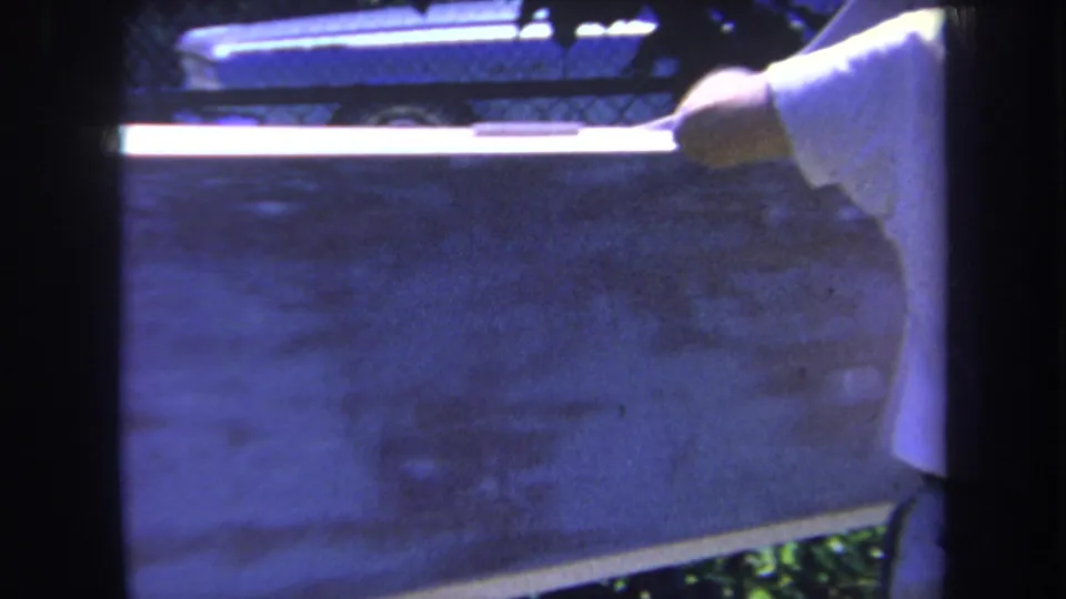 The man’s arm extends over a large pale surface, with a chain-link fence and dim outdoor background visible in the 8mm frame.
