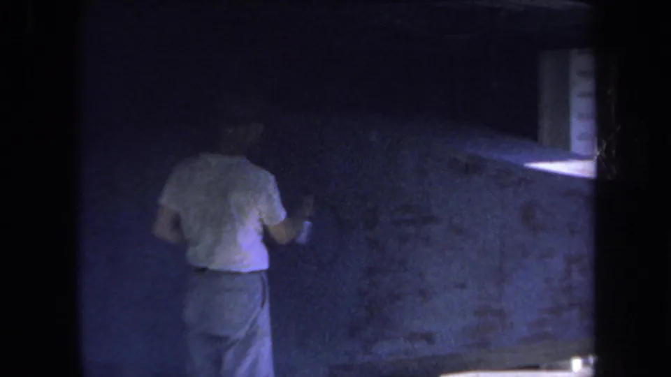 A wider 1969 8mm view shows the man standing near the large surface, with a parked car and shadowed surroundings in the background.