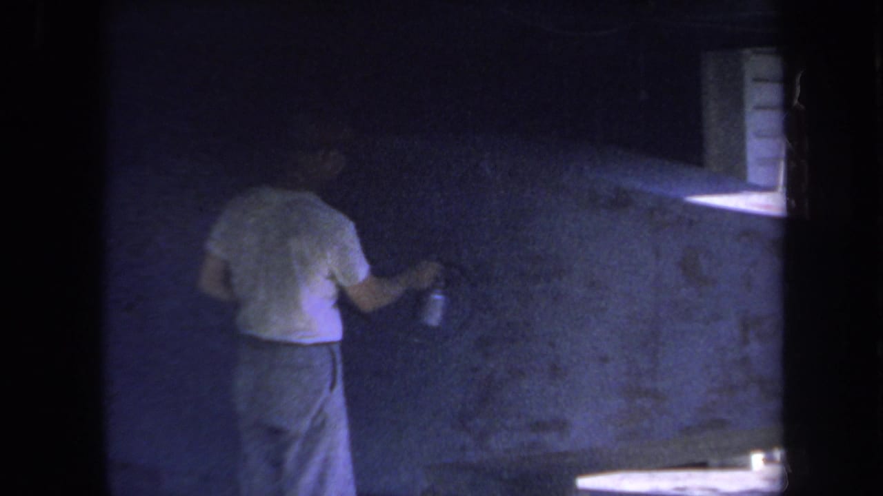 1969 Yucaipa home-movie frame showing a man painting a blue wall with a roller in a dim indoor space.