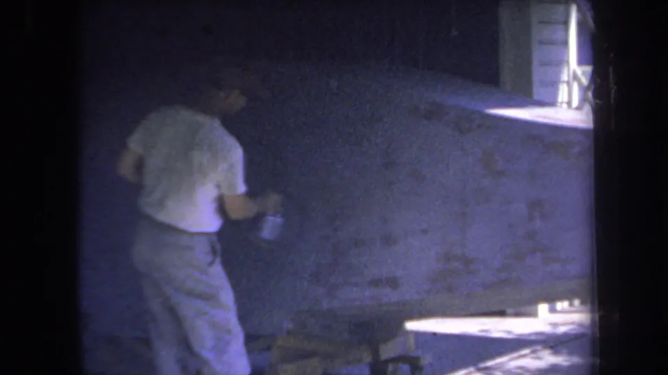 1969 Yucaipa home-movie still showing a man rolling paint on a blue wall in a shadowed interior room.