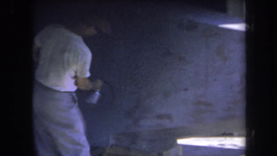 1969 Yucaipa frame of a man painting a blue interior wall, with a bright ledge or work surface at lower right.