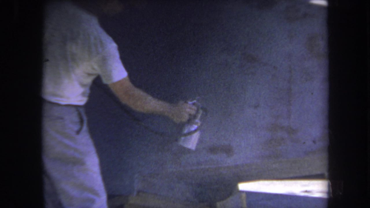 Man in a white T-shirt sprays a wood surface inside a dim workshop.