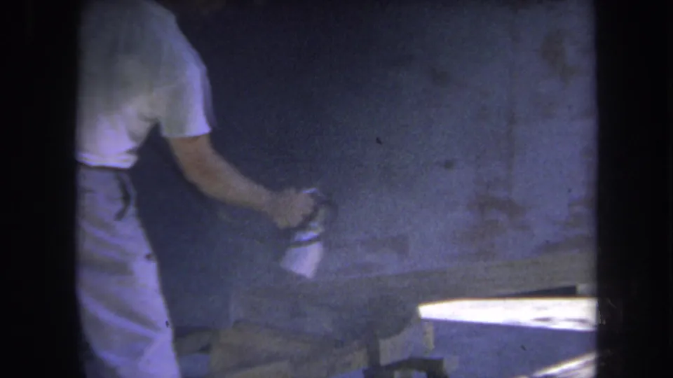 1969 Yucaipa home-movie frame showing a man using an aerosol can near wood in a dim workshop setting.