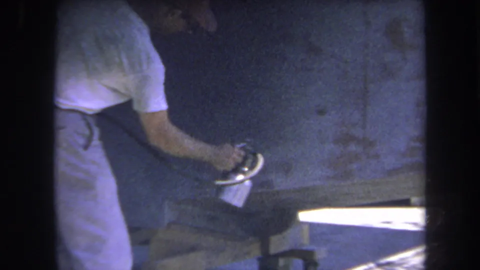 1969 8mm frame showing a man leaning over a wood surface while using a spray can in a private Yucaipa interior.