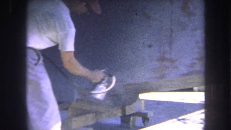 1969 Yucaipa 8mm frame of a man spraying a wooden board beside a bright tabletop or work surface in a workshop.