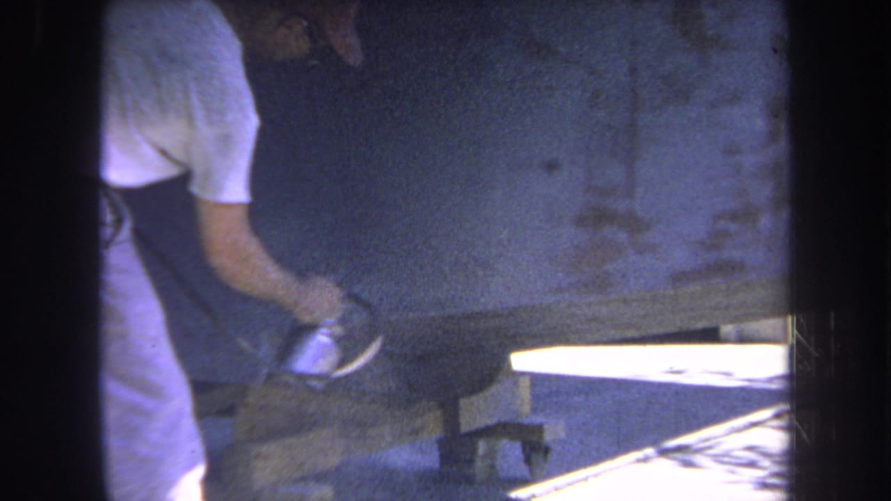Man in light clothing holds a spray gun near a large metal surface in sunlight.