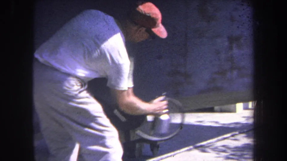 Worker in a red cap bends over with a spray gun while coating or preparing a large metal surface in bright outdoor light.