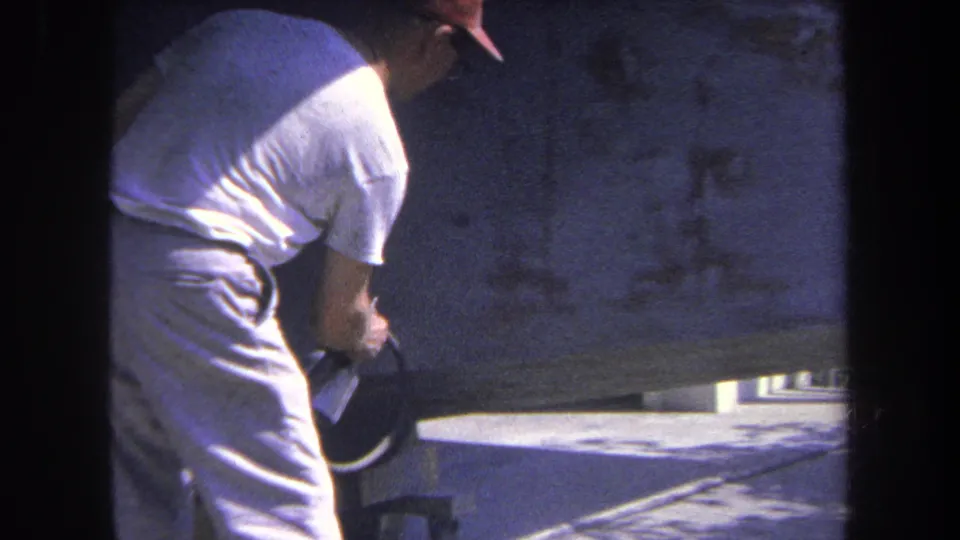 8mm frame of the worker's torso and arm as he handles the spray gun near the lower edge of a large metal surface.