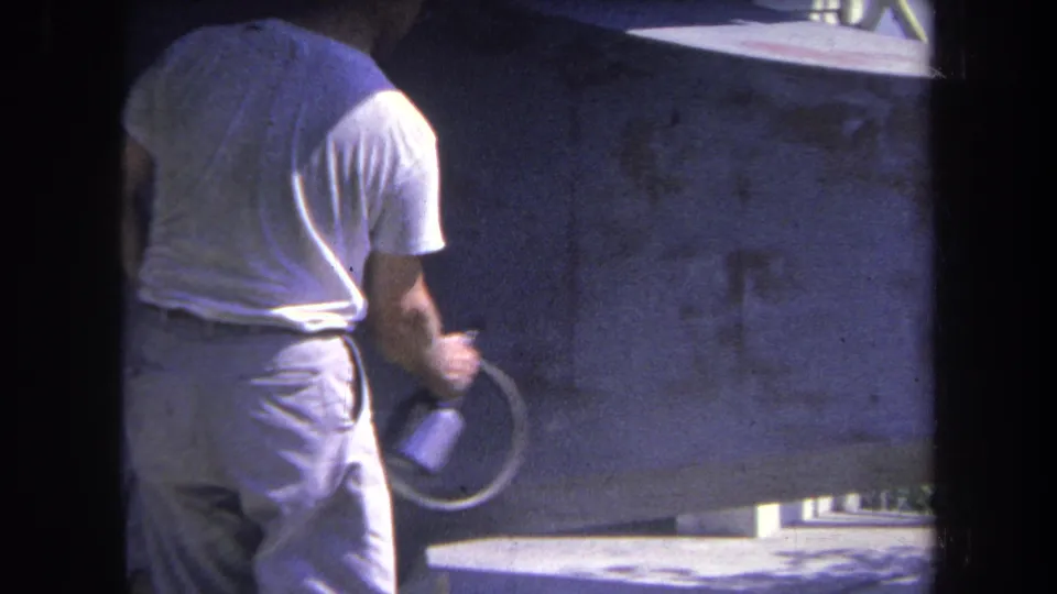 Worker seen from behind in 1969 California footage, holding a hose-fed spray gun beside a large metal panel.
