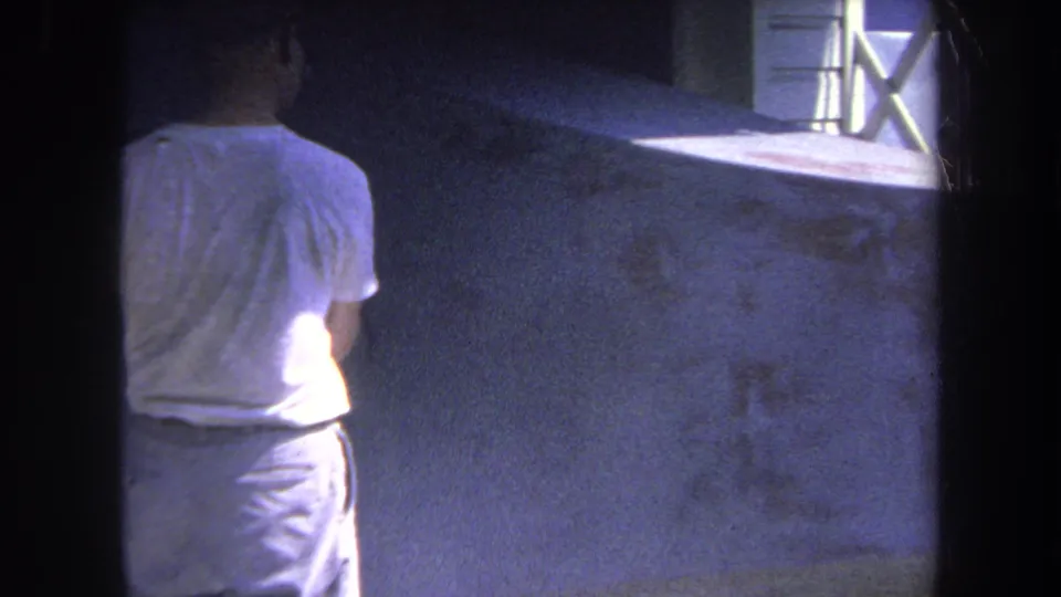 Faded 1969 8mm frame showing the worker partly in shadow beside a broad concrete wall in Yucaipa, California.