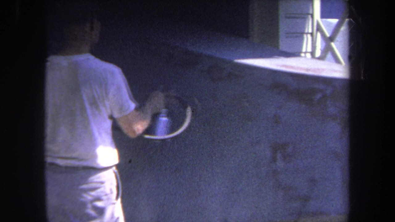 1969 8mm frame from Volmer Yucaipa, California, showing a man from behind using a spray gun on a dark wall beside a white-framed window.