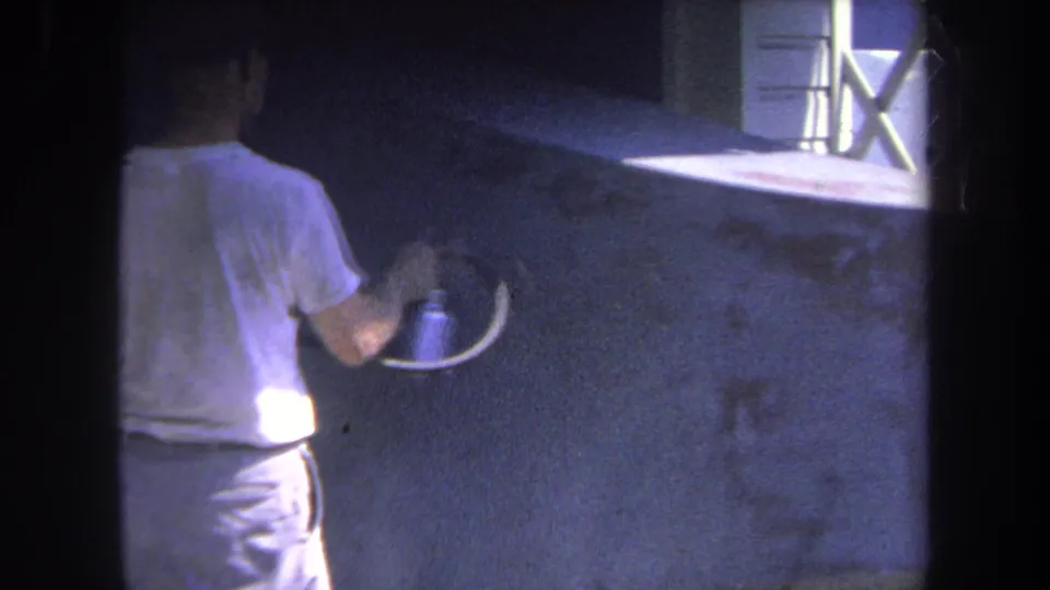 1969 8mm frame from Volmer Yucaipa, California, showing a man from behind using a spray gun on a dark wall beside a white-framed window.