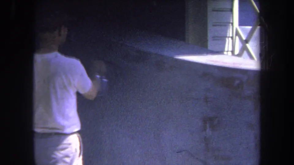 1969 Volmer Yucaipa home-movie frame showing the man applying spray paint across a dark wall, with a bright cross-braced window at right.