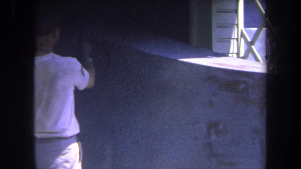 Volmer Yucaipa 1969 8mm frame with a man in a white shirt standing close to a wall, spray gun raised near a sunlit window opening.