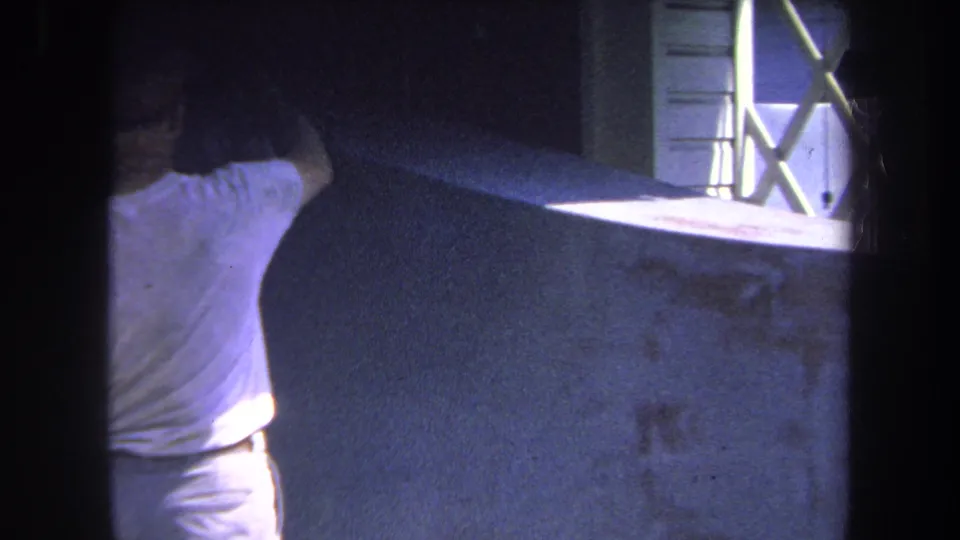 1969 8mm home-movie frame showing the man reaching higher with the spray gun while painting the wall near a white-framed window in Volmer Yucaipa.