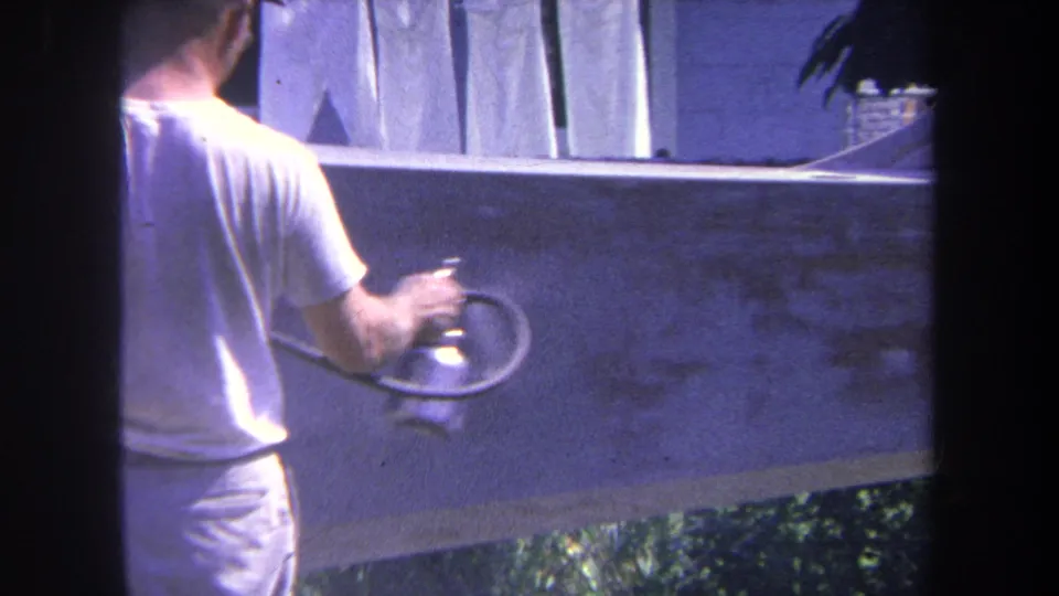 A close home-movie view shows the man holding the spray gun near the concrete wall during the outdoor painting task.