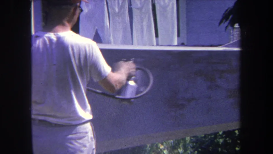1969 Yucaipa-area 8mm frame showing a man from behind using a spray gun on a backyard wall with laundry above.