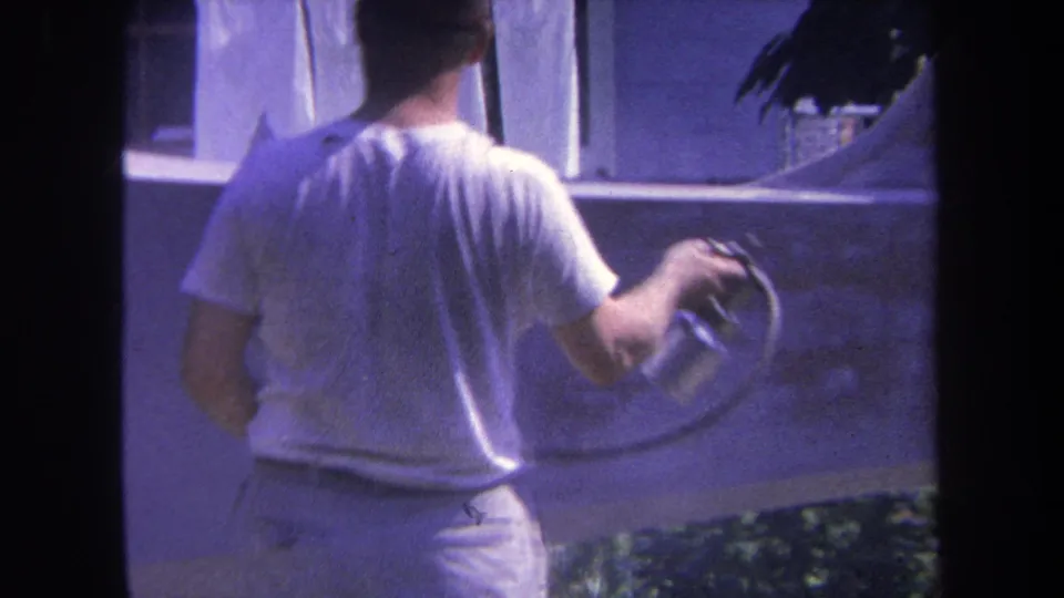 8mm frame from 1969 showing a man seen from behind continuing to spray paint a backyard wall in Yucaipa-area footage.