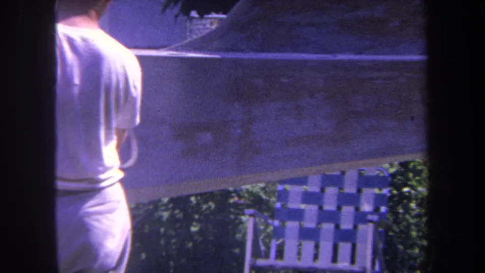 1969 home-movie frame showing the man partly turned away near the painted wall, shrubs, and a blue and white striped lawn chair.