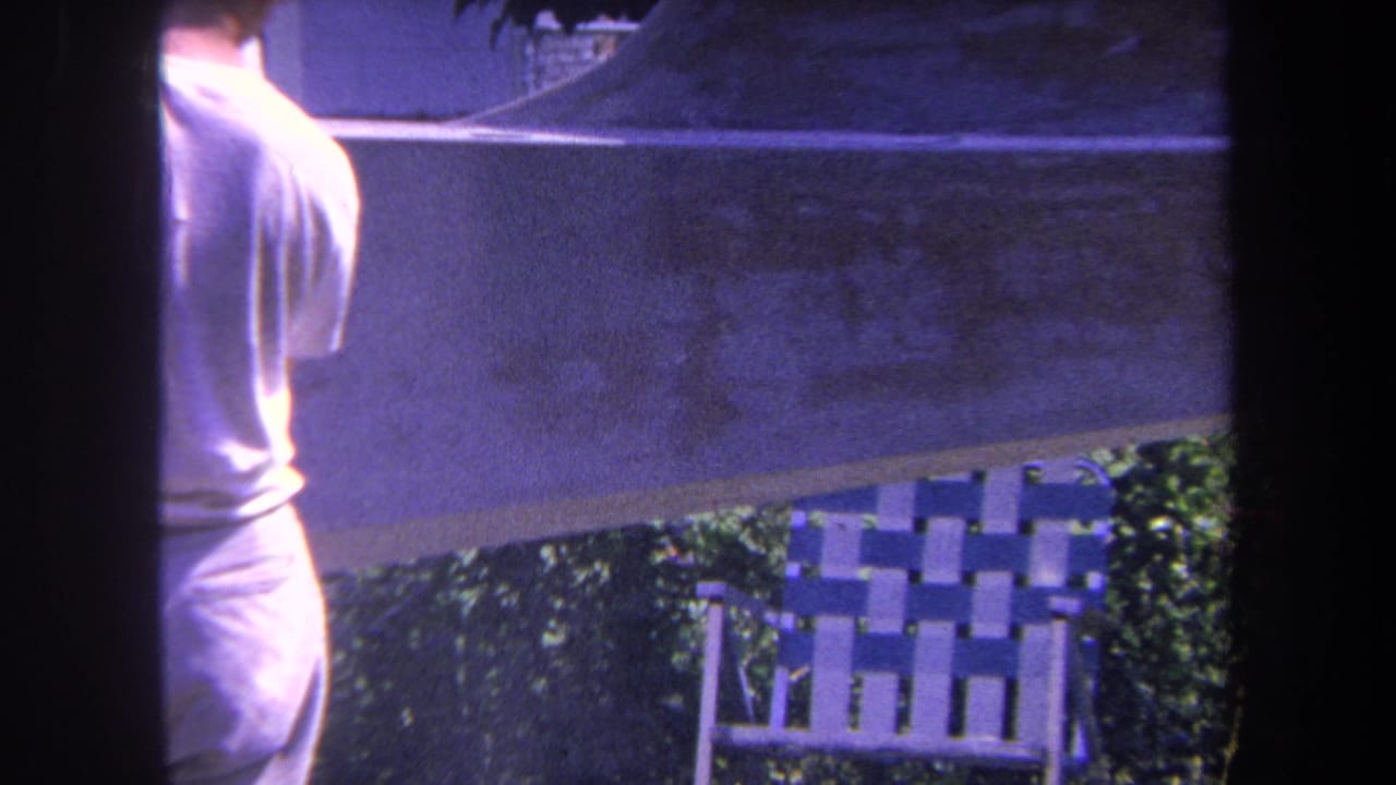1969 Yucaipa-area 8mm frame showing a man beside an outdoor surface, with a woven patio chair and greenery in the background.
