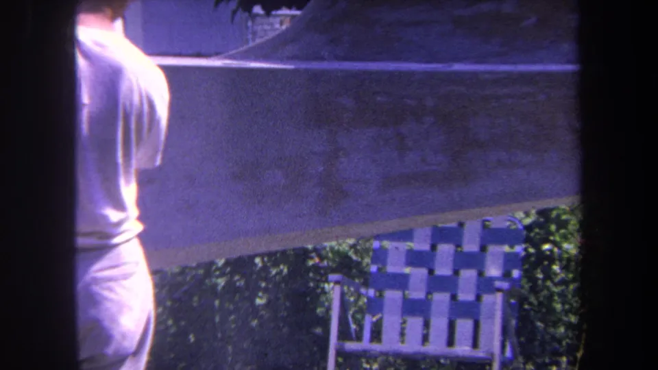 1969 Yucaipa-area 8mm frame showing a man beside an outdoor surface, with a woven patio chair and greenery in the background.