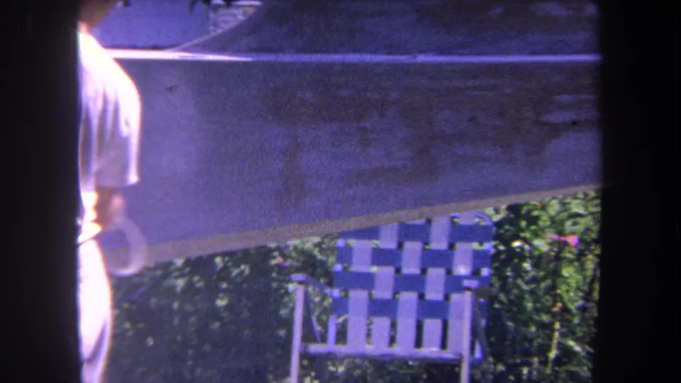 Soft 8mm frame showing the man partially cut off at the left as the outdoor surface, chair, and greenery fill the scene.