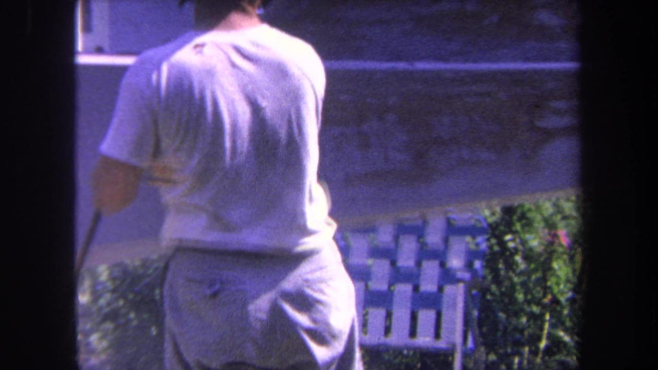 Adult man in a white T-shirt and light pants works beside a boat hull in 1969 Yucaipa backyard footage.