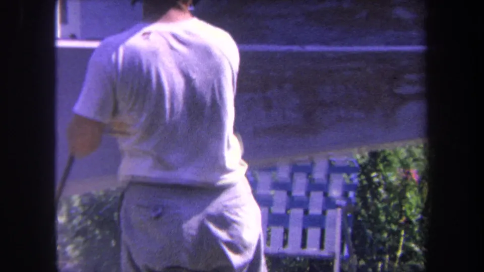 Adult man in a white T-shirt and light pants works beside a boat hull in 1969 Yucaipa backyard footage.