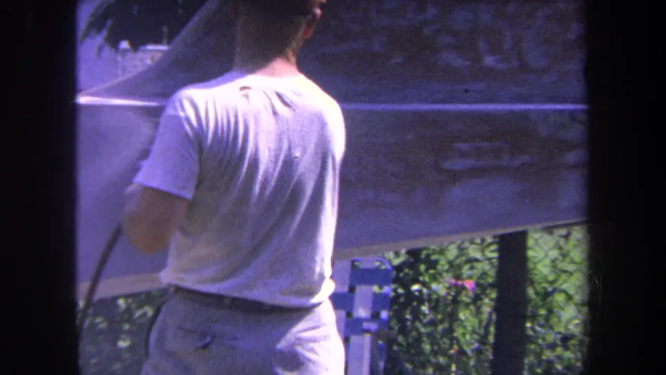In 1969 Yucaipa footage, a man is shown from behind holding a spray hose beside a boat hull outdoors.