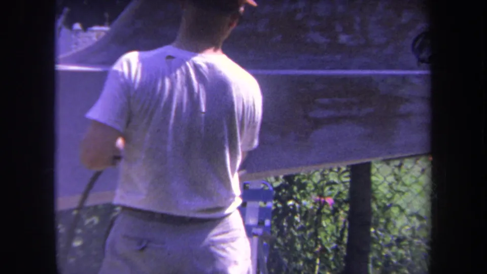 An adult man in light clothing faces the boat hull while holding a spray hose in vintage 8mm footage from Yucaipa.