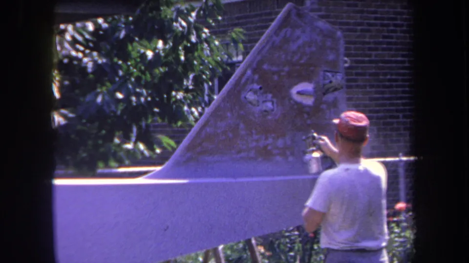 The 8mm frame shows the man spraying a weathered aircraft tail section, with greenery and a brick wall behind the work area.