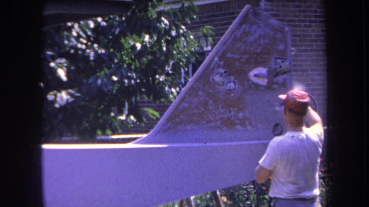 In 1969 Yucaipa footage, a man in a white shirt and red cap works on a weathered aircraft tail fin outside.